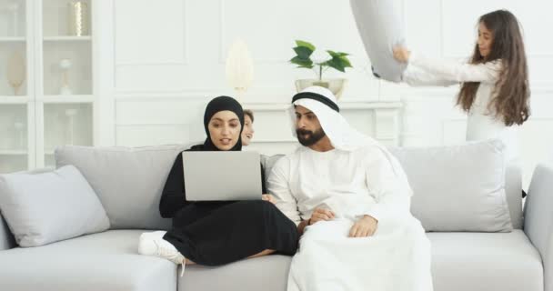 Arabische ouders in traditionele outfits zitten op de bank en werken aan laptop computer. Rumoerige kinderen rennen rond en vechten met kussens. Moeder en vader prediken kinderen, vertellen zich goed te gedragen — Stockvideo