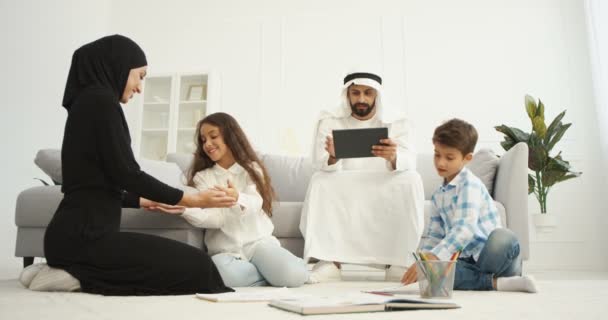 Αραβική μητέρα σε abaya φόρεμα παίζει το παιχνίδι με τα χέρια μικρό χαριτωμένο κόρη. Πατέρας στο dishdasha κάθεται στον καναπέ και χρησιμοποιώντας συσκευή tablet. Ο μικρός γιος στο πάτωμα ζωγραφίζει και ζωγραφίζει. Αραβική οικογένεια — Αρχείο Βίντεο