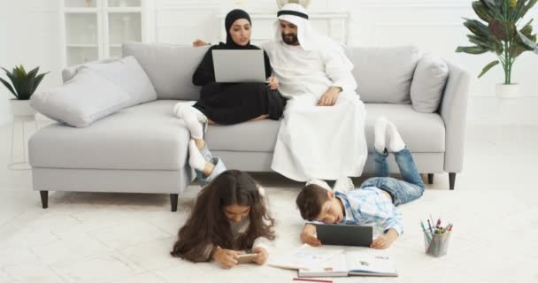 Arabian niños felices acostados en el suelo y jugando en gadgets. Hermana pequeña y hermano usando smartphone y tableta. Un par de padres sentados en el sofá con computadora portátil. Árabes madre y padre resto. — Vídeo de stock