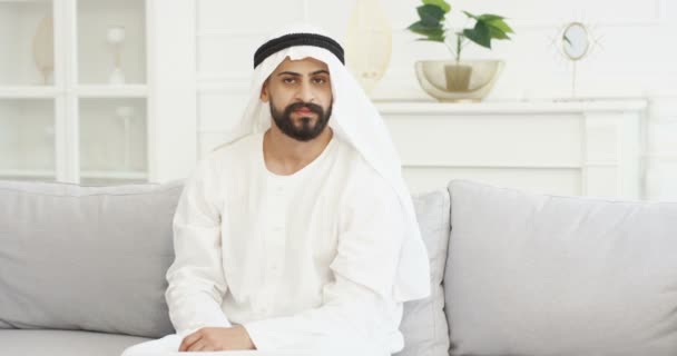 Retrato de belo jovem árabe em kandura sentado no sofá na sala de estar e sorrindo para a câmera. Muslim árabe macho em dishdasha no sofá em casa. Interior branco milionário árabe do Oriente Médio — Vídeo de Stock