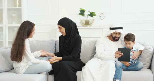 Keluarga Arab dengan anak-anak menghabiskan waktu bersama di ruang tamu. Muslim ibu dalam jilbab bermain tangan dengan putri kecil. Ayah Arab dengan anak laki-laki menggunakan perangkat tablet dan bermain. — Stok Video
