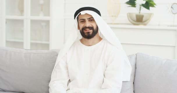 Portret przystojnego młodego araba w blogerze Kandura, siedzącego na kanapie w pokoju i rozmawiającego z kamerą. Mężczyzna vlogger mówi coś i wyjaśnia. Blogi wideo. Vlog z domu. Bliski Wschód. — Wideo stockowe