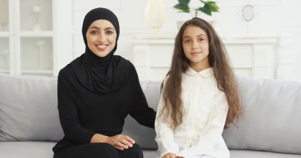 Retrato de una hermosa joven árabe en hiyab negro mirando a su linda hija adolescente con amor. Alegre encantadora madre musulmana en pañuelo para la cabeza y niña sonriendo a la cámara. Sentado en el sofá. — Vídeos de Stock