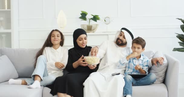 Weseli arabscy rodzice z małymi dziećmi siedzącymi na kanapie, uśmiechniętymi, oglądającymi filmy w telewizji i jedzącymi popcorn. słodkie arabskie małe dzieci z matka i ojciec spędzają czas razem w domu. — Wideo stockowe