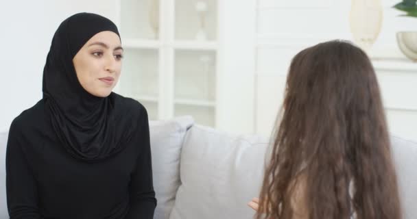 Bela jovem mãe em hijab conversando com a pequena filha adolescente. Mulher muçulmana em lenço de cabeça conversando com a menina no sofá em casa e abraçando. As mães adoram abraços. Traseira. Visão traseira . — Vídeo de Stock