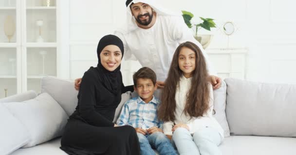 Retrato de familia árabe alegre con dos niños sentados en el sofá y sonriendo alegremente a la cámara. Pequeño lindo niño y niña con la madre y el padre en casa. Padres musulmanes con niños pequeños abrazándose. — Vídeos de Stock