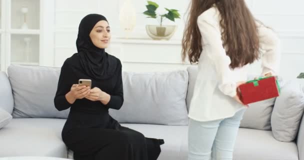 Dotter kommer till muslimsk mamma i hijab med närvarande bakom ryggen. Arabiska kvinna i Abaya klänning sitter på soffan och flicka lämnar sin gåva i rutan. Presentera för semester, födelsedag eller mors dag. — Stockvideo