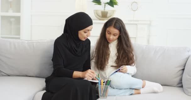 Moslim jonge moeder in zwarte hijab en abaya jurk tekening en kleurplaat met kleine tienerdochter op de bank in de woonkamer. Arabische vrouw en klein meisje samen schilderen tijdens het spelen thuis. — Stockvideo