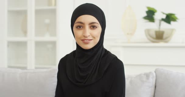 Retrato de la hermosa mujer joven árabe en vestido Abaya negro y gafas sentadas en el sofá y sonriendo a la cámara. Hermosa mujer musulmana guapa en hijab con sonrisa en el sofá en casa. — Vídeo de stock