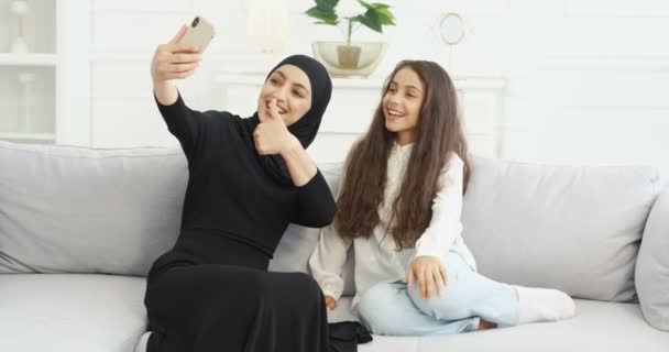 Beautiful young Arabian mother in hijab sitting on couch wih cute little daughter and taking selfie photos with smartphone camera. At home. Woman with kid making pictures selfies with mobile phone. — Stock Video