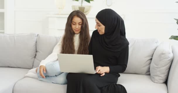 Muslim mother in headscarf talking with cute teen daughter, sitting on sofa and using laptop in living room. Pretty Arabian woman in hijab watching video on computer and browsing with teenage girl. — Stock Video