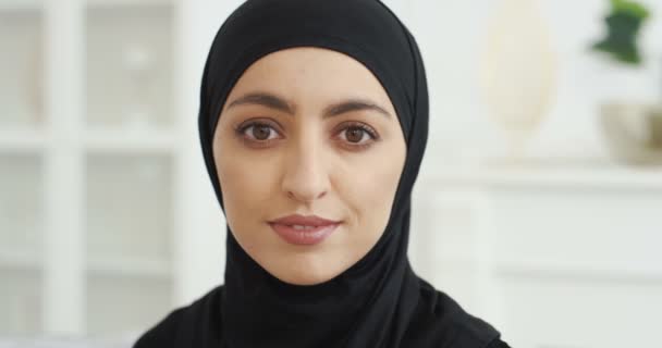 Retrato de una hermosa joven árabe musulmana en negro hijab tradicional mirando directamente a la cámara y sonriendo. En casa. Primer plano de cara bonita femenina con sonrisa. Pañuelo árabe. De interior. — Vídeo de stock