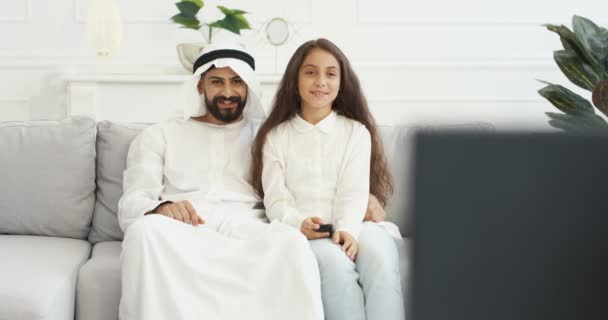 Arabo giovane padre bello seduto sul divano con la piccola figlia carina adolescente e guardare la TV. A casa. Piccola adolescente che cambia canale con telecomando e guarda la televisione con papà in una stanza accogliente — Video Stock