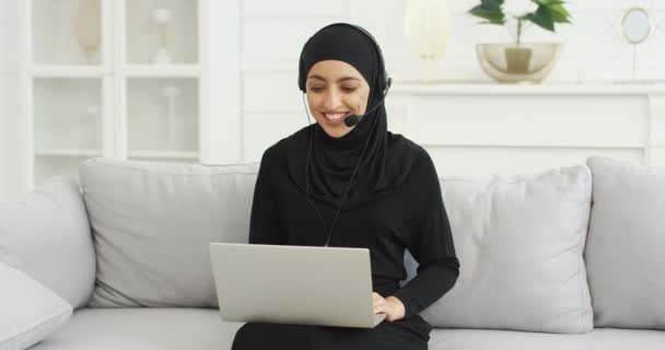 Pretty young Arabian woman blogger in headset sitting at laptop computer on couch and talking. Female vlogger in hijab working and educating. Video blogging. Home vlog studying. Muslim. Education. — Stock Video