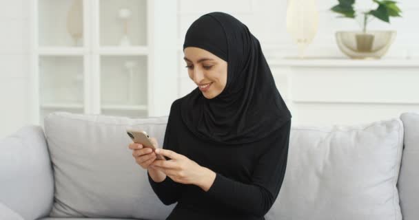 Cheerful young Arabian beautiful woman in black hijab sitting on couch and texting message on smartphone at home. Pretty smiled female muslim in headscarf tapping and scrolling on mobile phone. — Stock Video