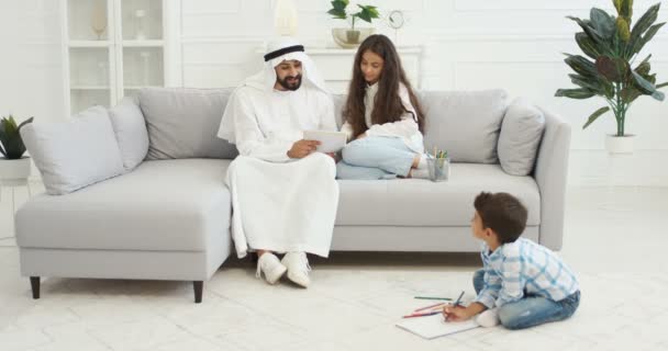 Arabisk far i vit kandura leker med små barn, dotter och son i vardagsrummet. Muselman pappa rita och måla bilder. Pojke och flicka med pappa målar hemma och umgås. — Stockvideo