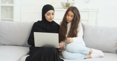 Oturma odasındaki kanepede oturan siyah tesettürlü genç Müslüman anne dizüstü bilgisayar kullanıyor ve konuşuyor. Küçük bir kızla güzel bir Arap kadın internette geziniyor..