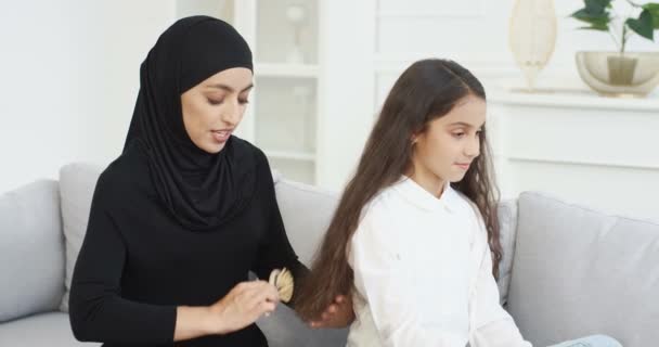 Молодая красивая арабская женщина в черном хиджабе улыбается и расчесывает длинные волосы довольно маленькой девочки-подростка с расчёской. Дома на диване. Мусульманская мать делает прическу для маленькой дочери-подростка в номере. — стоковое видео