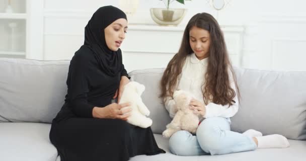 Mãe jovem muçulmana em vestido Abaya e hijab preto brincando com a filha pequena adolescente bonito com ursinhos de pelúcia. Mulher árabe em lenço de cabeça e menina brincar com brinquedos no sofá em casa . — Vídeo de Stock