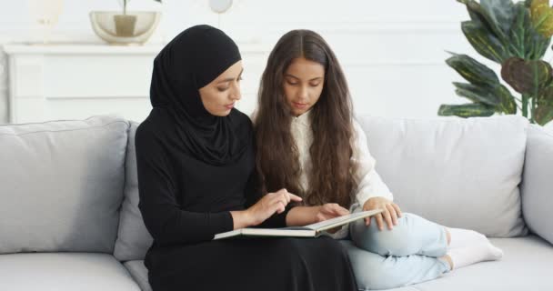Молодая арабская женщина в черном платке читает интересную историю в книге для довольно маленькой девочки-подростка. Дома на диване. Мусульманская мать обучает и воспитывает маленькую дочь-подростка с помощью учебника. — стоковое видео