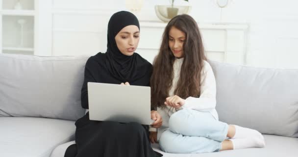 Musulmana joven madre en negro hijab sentado en el sofá en la sala de estar con linda hija adolescente utilizando el ordenador portátil y hablando. Mujer árabe bonita con pequeña adolescente navegando en línea en el ordenador. — Vídeo de stock