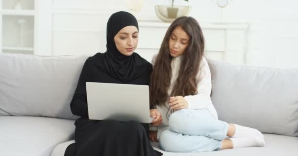 Mãe jovem muçulmana em hijab preto sentado no sofá na sala de estar com a filha adolescente pequena bonito usando laptop e conversando. Mulher muito árabe com menina adolescente navegando on-line no computador . — Vídeo de Stock