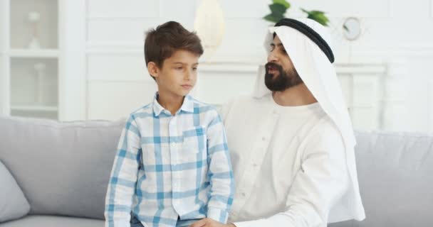 Portrét mladého pohledného muslimského otce v kanduře sedícího na gauči v obývacím pokoji s roztomilým malým synem, jak spolu mluví, dává si pět a usmívá se na kameru. Táta s malým klukem na gauči doma. — Stock video