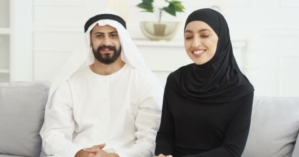 Portret van Arabieren moslim gelukkig paar zwaaiend met handen naar camera en lachen. Man en vrouw Arabieren zitten thuis op de bank en glimlachen. Midden-Oosten. Man in kandura en vrouw in hijab. — Stockvideo