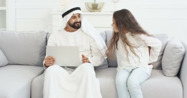 Daughter coming to muslim father in kandura with laptop and holding present behind back. Arabian man sitting on couch and girl handing him gift in box. Presenting for holiday, birthday or Fathers day — Stock Video