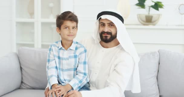 Portrait de jeune beau père musulman dans dishdasha assis sur le canapé dans le salon avec mignon petit fils sur les tours, se regardant et souriant à la caméra Happy papa avec petit garçon sur le canapé à la maison — Video