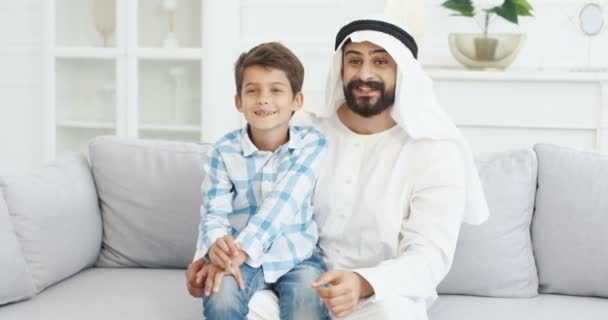 Retrato de jovem bonito pai muçulmano em kandura sentado no sofá na sala de estar com o filho pequeno bonito em voltas, falando, acenando com as mãos e sorrindo para a câmera. Pai com menino no sofá em casa . — Vídeo de Stock