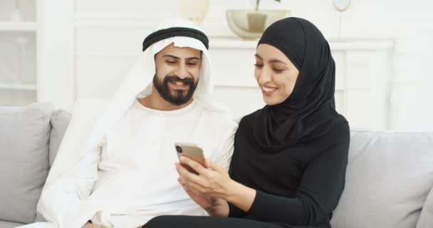 Ein junges arabisches, fröhliches Paar sitzt auf der Couch und schaut sich etwas auf dem Smartphone-Bildschirm an. Zu Hause. Schöner Mann im Kandura, schöne Frau im Hijab mit Handy, tippen und scrollen. Araber. — Stockvideo
