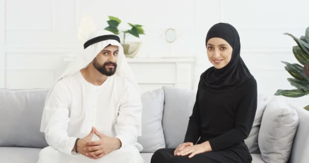 Arap müslüman genç çiftin portresi geleneksel Ortadoğu kıyafetleriyle evdeki kanepede oturuyorlar. Kanduralı bir Arap ve tesettürlü bir kadın aşkla bakıyor ve kanepede kameraya gülümsüyor.. — Stok video