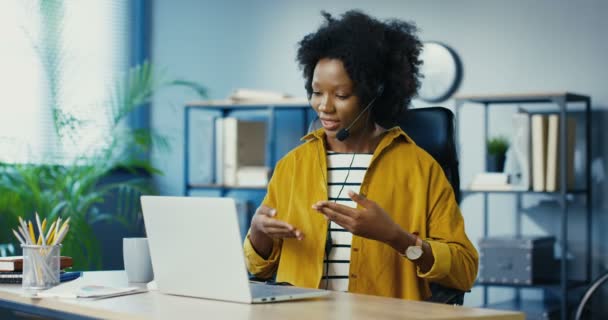 Ziemlich glückliche junge Afroamerikanerin sitzt im Büro und unterhält sich per Webcam auf dem Laptop mit Headset. Schöne Arbeiterin mit Videoanruf am Computer am Arbeitsplatz in Innenräumen. Jobkonzept — Stockvideo