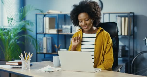 Trabajadora bastante femenina teniendo conversación de vídeo en la computadora en la oficina en el interior. Hermosa chica afroamericana alegre sentado en el escritorio y agitando la mano mientras habla en la webcam en el ordenador portátil de buen humor. — Vídeos de Stock