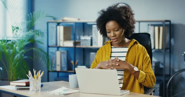 Hermosa mujer joven afroamericana alegre sentado en la oficina y hablando por webcam en el ordenador portátil de buen humor. Trabajadora bastante femenina teniendo video conversación en la computadora en el lugar de trabajo. Concepto de trabajo — Vídeo de stock