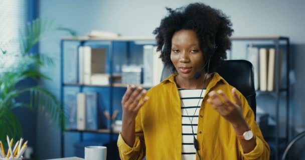 Porträt einer schönen, fröhlichen Arbeiterin, die am Arbeitsplatz im Haus etwas erklärt und Handgesten macht. Ziemlich glückliche Afroamerikanerin, die im Büro sitzt und mit Headset spricht. Jobkonzept — Stockvideo