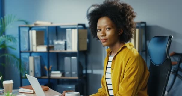 En söt afroamerikansk tjej ler mot kameran när hon sitter vid skrivbordet på kontoret inomhus. Porträtt av vacker glad lockigt ung kvinna på gott humör med leende på läpparna i skåpet. Känslor koncept — Stockvideo