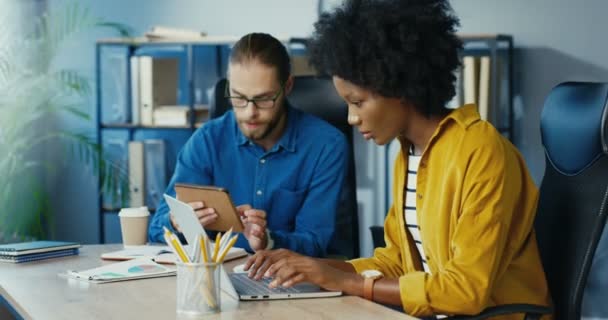 Travailleurs de bureau multiethniques à l'intérieur. Belle femme afro-américaine tapant sur ordinateur portable dans le cabinet. Joyeux homme blanc beau tapotant sur la tablette et parlant à une collègue féminine. Concept de bureau — Video