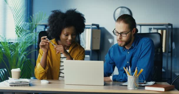 Multietniska yrkesverksamma som arbetar på bärbar dator på kontoret och chatta. Afroamerikansk tjej pratar med en man när hon sitter vid skrivbordet. Stilig vit man i glasögon som förklarar för kvinnlig kollega — Stockvideo