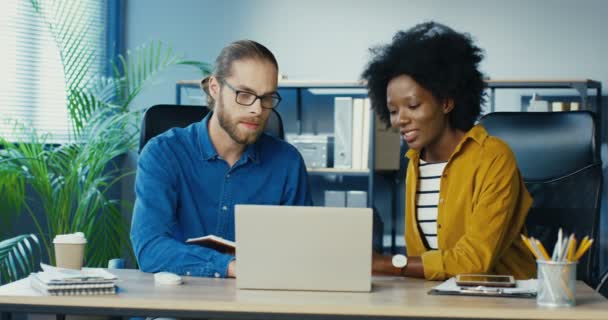 Ofiste dizüstü bilgisayarda çalışan ve sohbet eden melez ofis çalışanları. Güzel Afrikalı Amerikalı kız bir erkekle konuşuyor. Gözlüklü yakışıklı beyaz adam bayan meslektaşımla konuşuyor. — Stok video