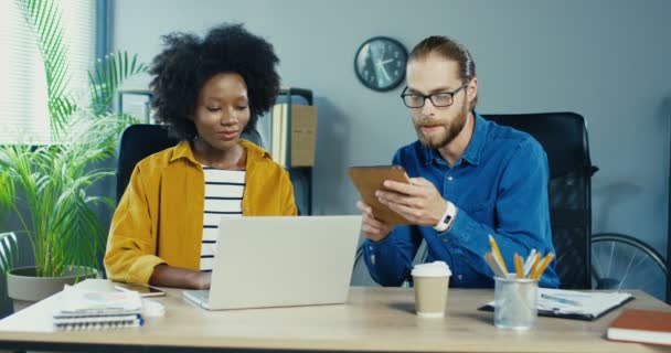 En vacker afroamerikansk kvinna som skriver på laptop i skåpet. Upptagen kaukasiska stilig man knacka på tablett och prata med kvinnlig kollega. Multietniskt kontorspersonal som arbetar inomhus. Kontorsbegreppet — Stockvideo