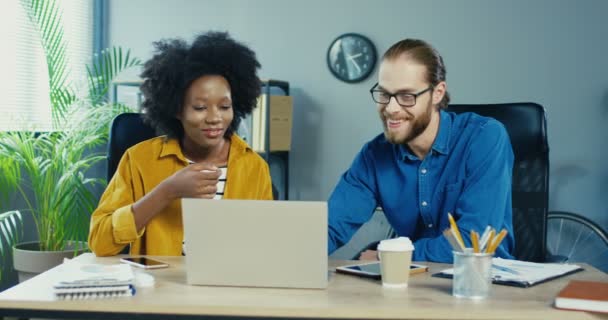 Mujer afroamericana y hombre caucásico trabajando en la computadora mientras está sentado en el gabinete. Hermosa chica mirando la pantalla y hablando con el hombre. Hombre guapo en gafas escribiendo en el portátil. Concepto de trabajo — Vídeos de Stock