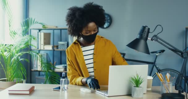 Hermosa mujer afroamericana en máscara y guantes de protección de limpieza portátil con antiséptico mientras está sentado en el lugar de trabajo. Trabajadora de oficina desinfecta computadora en gabinete en cuarentena en interiores — Vídeo de stock