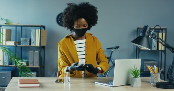 Γυναίκα υπάλληλος γραφείου απολύμανση ποντίκι υπολογιστή και smartphone σε καραντίνα. Όμορφη Αφροαμερικανή γυναίκα με μάσκα και προστατευτικά γάντια καθαρισμού κινητό τηλέφωνο με αντισηπτικό, ενώ κάθεται στην εργασία — Αρχείο Βίντεο