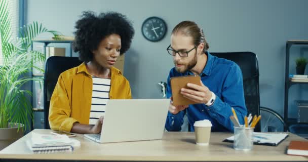 Jolie Afro-Américaine tapant sur un ordinateur portable au bureau. Occupé Caucasien bel homme tapotant sur tablette et parlant à une collègue féminine. Des gens de bureau multiethniques travaillant à l'intérieur. Concept d'emploi — Video