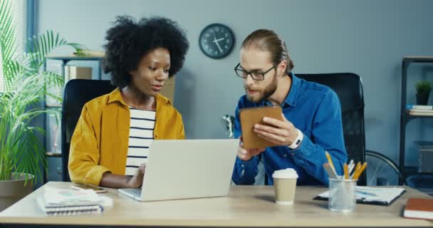 Mujer afroamericana bastante escribiendo en el portátil en la oficina. Hombre guapo caucásico ocupado tocando la tableta y hablando con una colega. Oficinas multiétnicas trabajando en interiores. Concepto de trabajo — Vídeo de stock