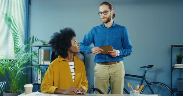 Piękna Afroamerykanka siedzi przy biurku w dobrym nastroju. Przystojny biały mężczyzna w okularach stojący z tabliczką. Pracownicy mieszanej rasy męskiej i żeńskiej patrzący sobie w oczy i uśmiechnięci do kamery — Wideo stockowe