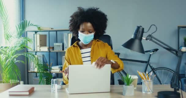 Schöne afroamerikanische Frau in medizinischer Maske, die Laptop mit antiseptischem reinigt, während sie am Schreibtisch am Arbeitsplatz sitzt. Hübsches Weibchen desinfiziert Computer im Schrank in Quarantäne drinnen. Bürokonzept — Stockvideo