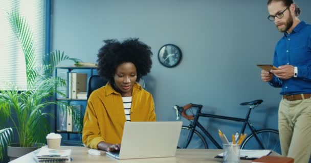 Pretty African American girl sitting at desk and typing on laptop. Caucasian man in glasses with tablet looking at computer screen while standing. Busy mixed-race male and female working at office. — Stock Video